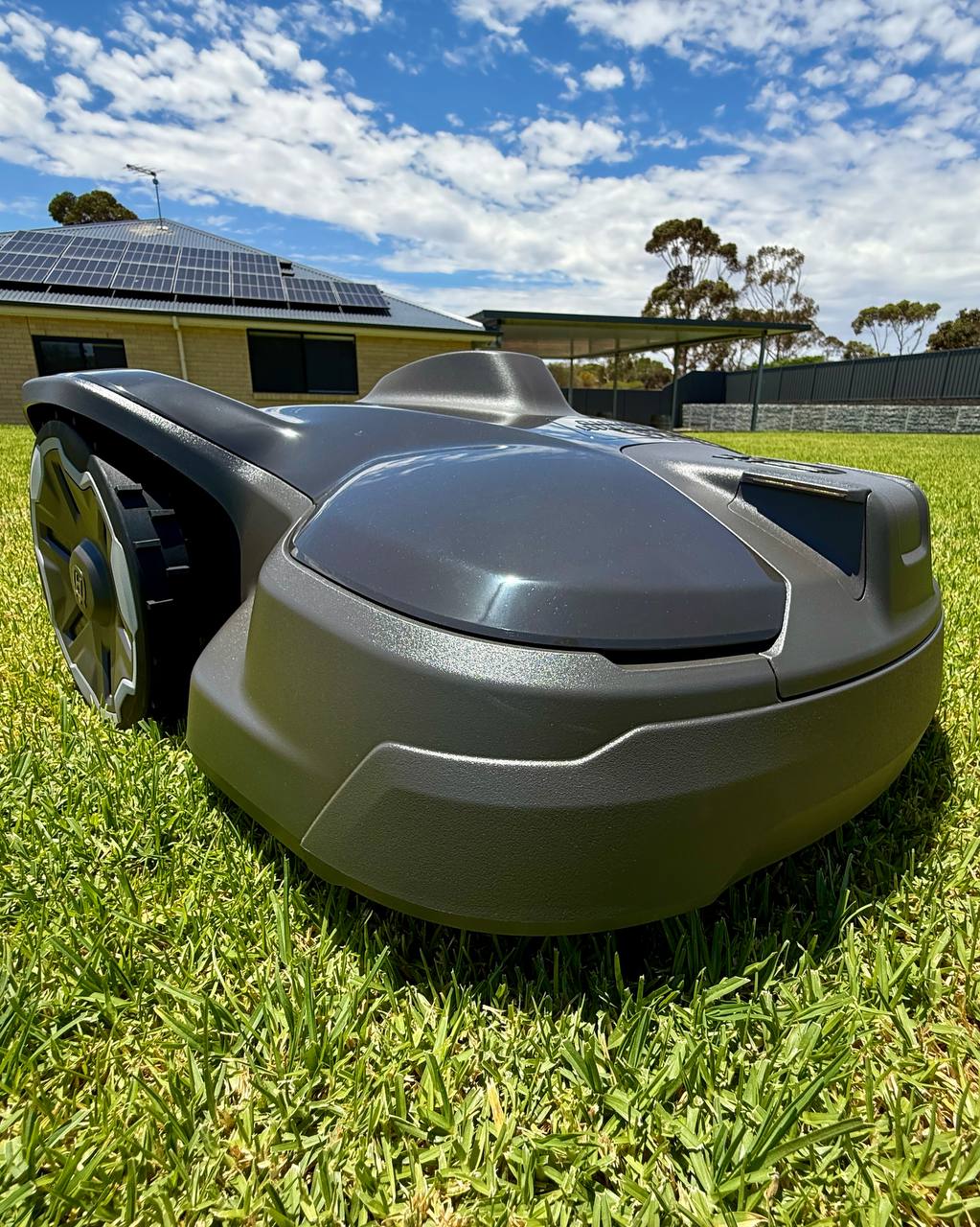 Husqvarna Automower 320 NERA on a grassy area with a house and solar panels in the background.