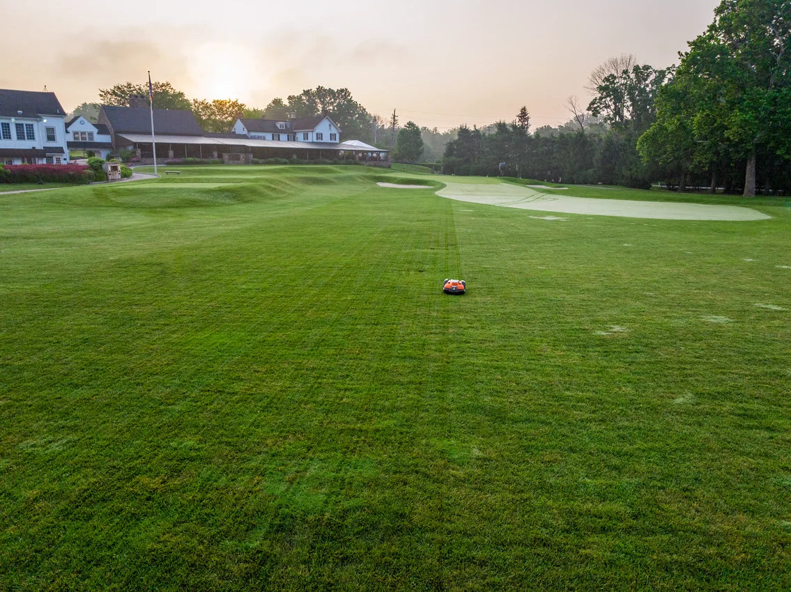 Golf course with an Automower 550 EPOS commercial robot lawn mower in the foreground, surrounded by trees and houses.